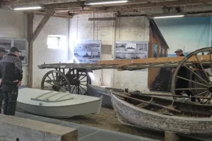 Museum interior displaying historical transport models: a wooden wagon with large wheels, a small rowboat, and visitors examining exhibits on brick walls and panels.