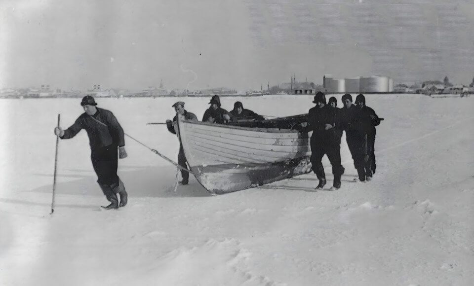 A group of men on a snow-covered landscape pull a small wooden boat along a rope toward a frozen shoreline with distant buildings in the background.
