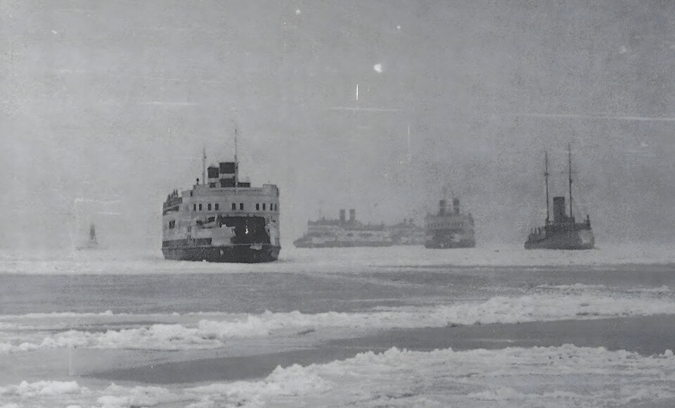Historic black-and-white photo of several ships on a frozen sea, with a large vessel in the foreground breaking through ice.