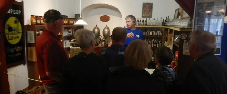 Small group of people listening to a man in a blue shirt giving a talk behind a bar with bottles on shelves.